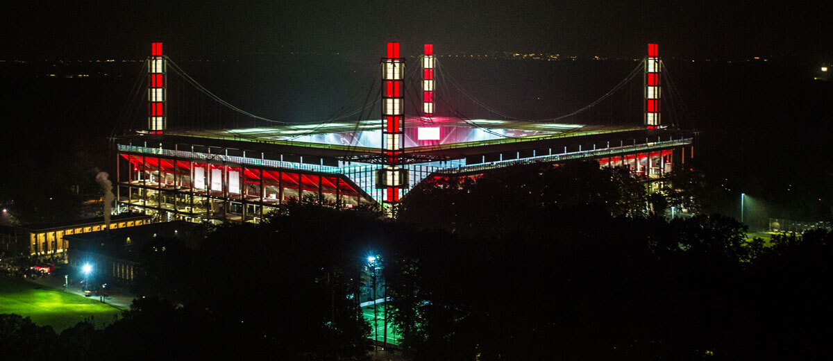 Rheinenergiestadion bei Nacht mit rot/weiß beleuchteten Pylonen